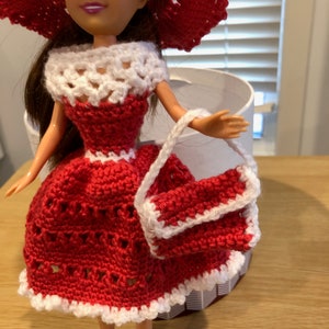 May include: A red crocheted dress with white trim and a matching red crocheted hat and handbag. The doll is wearing the outfit.