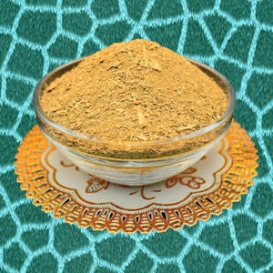 May include: A clear glass bowl filled with a mound of light brown, dried herbs or spices. The bowl sits on a decorative, round, gold-colored doily with a white center. The background is a teal fabric with a geometric pattern.