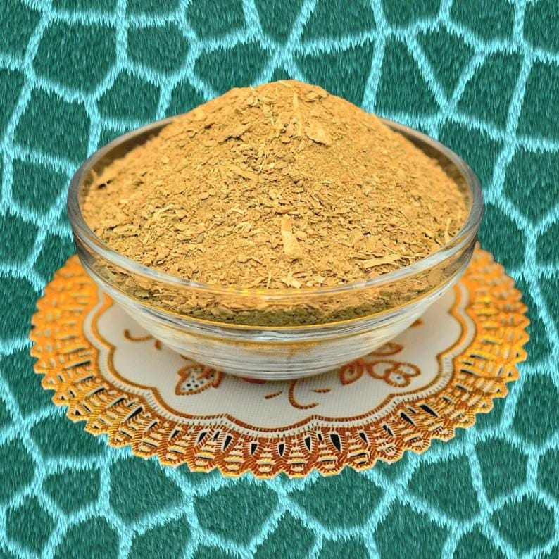 May include: A clear glass bowl filled with a mound of light brown, dried herbs or spices. The bowl sits on a decorative, round, gold-colored doily with a white center. The background is a teal fabric with a geometric pattern.