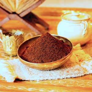 May include: A close-up of a small, round, gold-coloured bowl filled with dark brown coffee grounds. The bowl is sitting on a white, fringed cloth on a wooden surface.