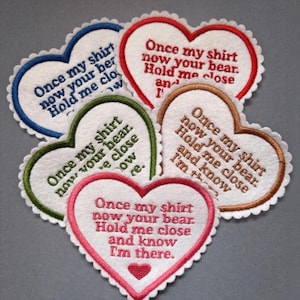May include: Five heart-shaped fabric patches with scalloped edges. Each patch features white fabric with embroidered text reading: Once my shirt now your bear. Hold me close and know I'm there. The embroidery colours are red, blue, green, brown, and pink.