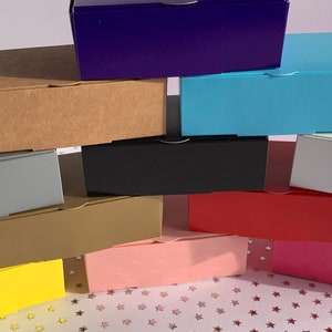 May include: A stack of ten small cardboard boxes in various colours, including purple, blue, brown, grey, black, gold, pink, and red. The boxes are arranged in a pyramid shape on a white surface with a star pattern.