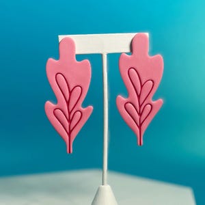 以下が含まれることがあります： 濃いピンク色の葉の形をしたピンク色のイヤリングのペア。 イヤリングは、青い背景に対して白いイヤリングスタンドに表示されます。 ユニークでスタイリッシュなアクセサリーです。