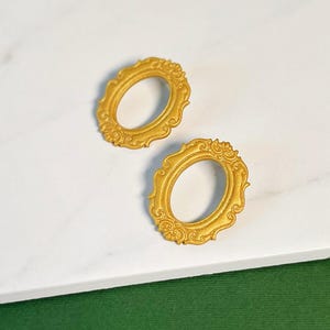 Puede incluir: Un par de pendientes ovalados de oro con un diseño ornamentado detallado. Los pendientes están hechos de un material metálico liso.