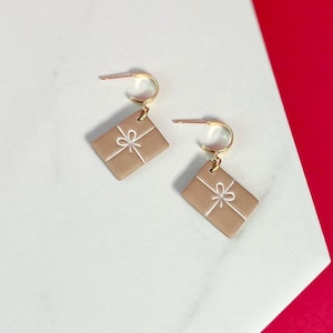May include: A pair of earrings featuring a tan rectangular shape with a white bow design. The earrings have gold-colored hoops. The background is white and red.