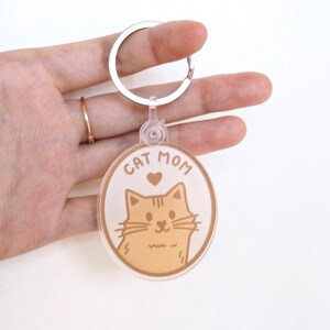 May include: A circular acrylic keychain featuring a light orange illustration of a cat face with whiskers and a small heart above it. The text CAT MOM is printed in a brown sans-serif font at the top. The keychain has a silver metal ring.