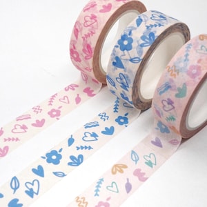May include: Three rolls of decorative adhesive tape featuring repeating patterns of hearts, flowers, and leaves. The first roll has pink motifs, the second roll displays blue designs, and the third roll showcases a multicolored print on a light background.