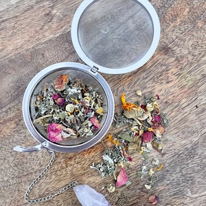 May include: A silver tea infuser ball filled with dried herbs, flowers, and rose petals. The infuser is open and the contents are spilling out onto a wooden surface. A purple amethyst crystal is attached to the infuser chain.