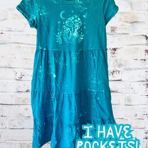May include: A turquoise blue tiered dress with a white mushroom and moon graphic. The dress has short sleeves and the text "I HAVE POCKETS!" is printed on the bottom.