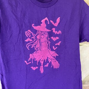 May include: A purple t-shirt featuring a pink witch design with a pointed hat, flowing hair, and a dress, surrounded by bats. The graphic is centered on the shirt, creating a Halloween theme.