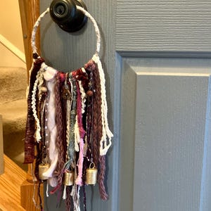 May include: A handcrafted door hanging with a circular top and dangling strands of yarn and fabric in shades of burgundy, white, and pink. Small gold bells are attached to the bottom of the strands, creating a bohemian aesthetic.