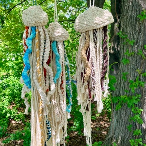 May include: Three hanging jellyfish decorations crafted from off-white macrame cord. Each features a rounded, knotted head and long, trailing tentacles in various textures and colors, including shades of blue, brown, and cream, suspended by loops.