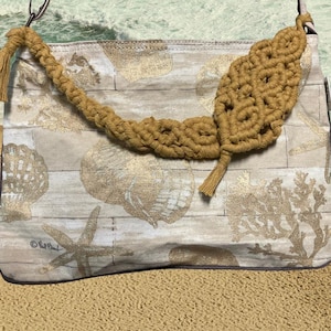 May include: A beige crossbody bag with a macrame strap and seashell print. The bag features a light brown macrame strap and a beige background with gold seashell, starfish, and coral designs. The bag has a dark blue trim and the text "OWLL" is visible.