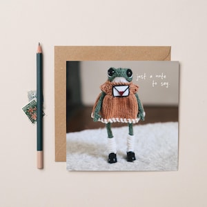 Op de afbeelding: Een wenskaart met een gebreide kikker die een jurk draagt en een envelop vasthoudt. De kikker is groen, bruin en wit, met de tekst "just a note to say". Een potlood, postzegels en een bruine envelop zijn ook aanwezig.