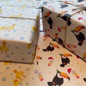 May include: Two rolls of wrapping paper, one with a duckling pattern and the other with a toucan pattern. Both rolls are white with a light blue background. The duckling pattern features yellow ducklings with blue bubbles. The toucan pattern features black and orange toucans with party hats and yellow stars.