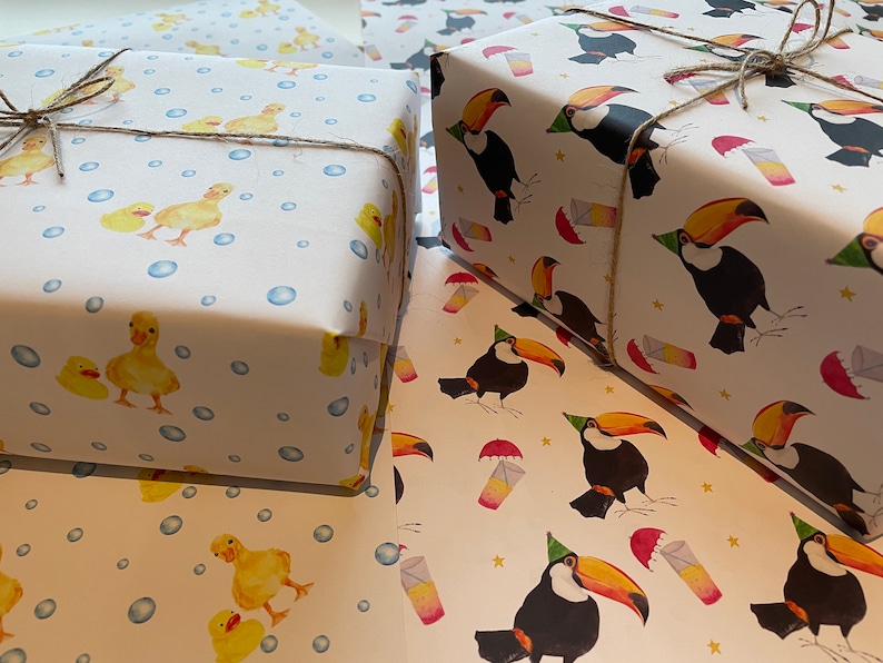 May include: Two rolls of wrapping paper, one with a duckling pattern and the other with a toucan pattern. Both rolls are white with a light blue background. The duckling pattern features yellow ducklings with blue bubbles. The toucan pattern features black and orange toucans with party hats and yellow stars.