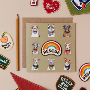 Puede incluir: Una tarjeta de felicitación con retratos de perros y parches bordados. Los parches muestran frases como "Best Buddy", "Rescue" y "Belly Rub Club". También hay un lápiz y un pañuelo rojo.