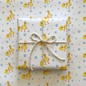 May include: White gift wrap with a repeating pattern of yellow rubber ducks and blue bubbles. The gift wrap is tied with a natural twine bow.