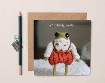 May include: A square greeting card featuring a knitted frog wearing a cream sweater with brown leaf patterns and orange pumpkin-style pants with brown suspenders. The text it's spooky season is printed above the frog. A dark pencil lies to the left.