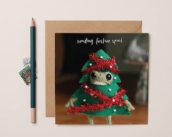 May include: A square greeting card featuring a green felt creature shaped like a Christmas tree, decorated with red tinsel and white beads, with the text "sending festive spirit" printed above it, accompanied by a dark pencil and a brown envelope.