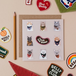 Könnte beinhalten: Eine Grußkarte mit Aquarellillustrationen von Katzen mit Bandanas. Um die Karte herum befinden sich gestickte Aufnäher mit Aufschriften wie "Best Buddy", "Team Toe Beans" und "Belly Rub Club". Ein Bleistift und ein rotes Scout's Honour-Bandana sind ebenfalls zu sehen.
