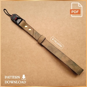 May include: A brown leather wrist strap with a black and red camera attachment. The strap has two gold-colored rivets and a loop for attaching to a camera. The image includes the text "PATTERN DOWNLOAD" and a PDF icon.