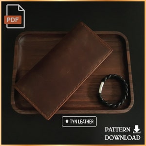 Op de afbeelding: Een bruine leren portemonnee op een houten dienblad met een zwarte leren armband. De tekst "TYN LEATHER" is zichtbaar op het dienblad. De tekst "PATTERN DOWNLOAD" is zichtbaar onder het dienblad.
