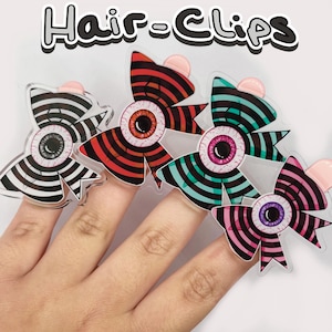May include: A hand displays four novelty hair clips. Each clip features a bow design with an eyeball center and striped patterns in black and white, red and black, teal and black, and pink and black. The text "Hair Clips" is at the top.