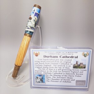 May include: Handcrafted pen with a light wood barrel and decorative metal accents. The pen features a detailed image of a scene with a building and a white owl. The pen is accompanied by a card that reads "Durham Cathedral".
