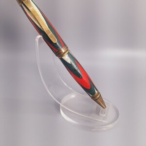 May include: A decorative pen with a red, green, and white patterned design. The pen has brass-colored accents and is displayed on a clear acrylic stand. The pen's design features a series of angled lines.