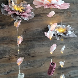 Könnte beinhalten: Dekorative Blumen-Mobile zum Aufhängen mit Papierblumen in Rosa, Grau und Weiß. Die Blumen werden von kleinen LED-Lichtern beleuchtet, die ein sanftes Leuchten erzeugen. Das Mobile enthält hängende Dekorationselemente.