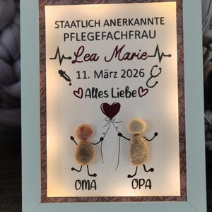 Könnte beinhalten: Gerahmtes Kunstwerk mit weißem Rand, das Text und Illustrationen zeigt. Der Text enthält einen Namen, ein Datum und deutsche Sätze. Zwei Kieselsteinfiguren halten einen Ballon. Der Rahmen hat einen warmen, beleuchteten Hintergrund.