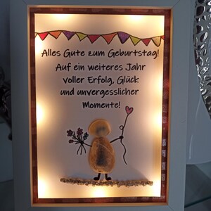 Könnte beinhalten: Ein gerahmtes Kunstwerk mit weißem Rahmen, das eine Kieselkunstfigur mit Blumen und einem herzförmigen Ballon zeigt. Das Kunstwerk enthält den deutschen Text "Alles Gute zum Geburtstag!" und wird von kleinen Lichtern beleuchtet.