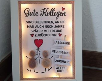 Può includere: Un'opera d'arte incorniciata con il testo tedesco "Gute Kollegen" e un messaggio toccante. L'opera presenta figure di ciottoli che tengono un palloncino rosso, con cartelli che indicano "Addio", "Nuovo inizio" e "Futuro".