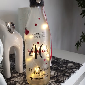 Könnte beinhalten: Eine dekorative Glasflasche mit Lichterketten, verziert mit roten Herzen und Text. Die Flasche ist mit einem grauen Stoffkragen, rot-weißem Band und einem silbernen Herzanhänger verziert. Der Text enthält ein Datum und Namen.