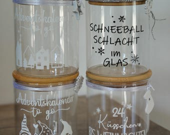 Könnte beinhalten: Vier durchsichtige Gläser mit Bambusdeckeln, verziert mit weißen oder schwarzen Vinylaufklebern. Die Designs zeigen Texte wie Adventskalender to go, Schneeballschlacht im Glas und 24 Küsschen bis Weihnachten sowie Grafiken von Häusern, Wichteln und Schneeflocken.