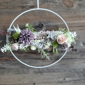 Könnte beinhalten: Ein weißer, kreisförmiger Metallring, geschmückt mit einem Blumenarrangement aus einer großen violetten Dahlie, einer blassrosa Rose, weißen Tannenzapfen, grünen Zweigen und einem silbernen Stern. Das Dekorationsstück hängt an einem weißen Band.
