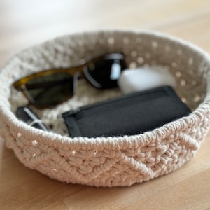 Puede incluir: Un cuenco de crochet blanco con un patrón tejido. El cuenco está lleno de varios artículos, incluyendo gafas de sol, una pequeña caja blanca y una cartera negra.