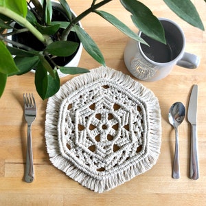 May include: A white crocheted placemat with a geometric design and fringe edges. The placemat is on a wooden table with a fork, spoon, and knife beside it.