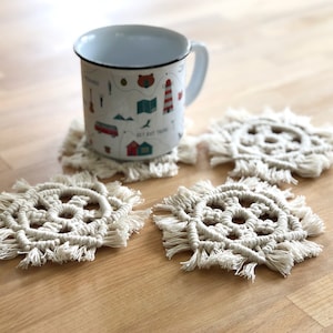Puede incluir: Cuatro posavasos de crochet blanco con detalles de flecos. Una taza de esmalte blanco con un diseño de mapa se encuentra sobre uno de los posavasos.