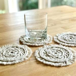 Puede incluir: Cuatro posavasos de crochet blanco con detalles de flecos, uno con un vaso encima. Los posavasos están sobre una superficie de madera.