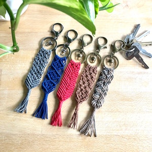May include: Five colourful macrame keychains with tassels. The keychains are made with different coloured cords and have a silver key ring attached.