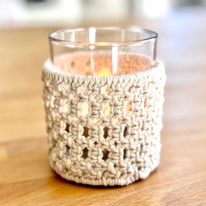 Op de afbeelding: Een glazen kaarsenhouder met een gehaakte beige hoes. De kaarsenhouder staat op een houten oppervlak.