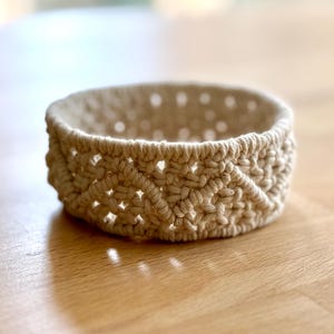 以下が含まれることがあります： 幾何学模様の入った、クリーム色の小さなマクラメボウル。ボウルはコットンコードで作られており、丸い形をしています。