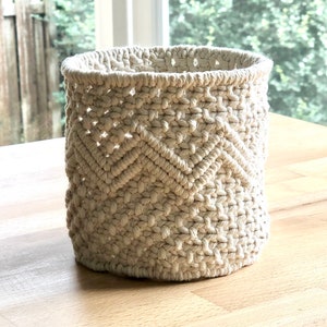 May include: A white macrame basket with a chevron pattern. The basket is made of natural cotton rope and has a round shape.