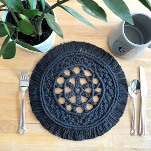 Puede incluir: Salvamanteles de crochet negro con borde de flecos y un diseño circular en el centro. El salvamanteles está sobre una mesa de madera con un tenedor, una cuchara y un cuchillo a su lado.
