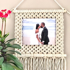 Puede incluir: Un tapiz de macramé blanco con una foto de una pareja besándose frente a una playa. La foto está enmarcada por el macramé y el tapiz tiene una franja en la parte inferior.
