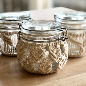 May include: Three glass jars with metal lids are wrapped in beige macrame. The jars are sitting on a light brown wooden surface.