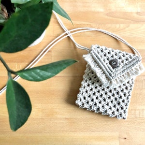 May include: A beige macrame phone bag with a button closure and fringe detail. The bag has a woven design and a long, thin strap. The bag is shown on a light brown wooden surface, with a green plant in the background.