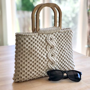Può includere: Una borsa tote in macramé color crema con manici in legno. La borsa ha un motivo geometrico ed è appoggiata su una superficie in legno con un paio di occhiali da sole neri.
